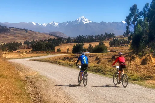 Two guests cycling down gravel road, golden grasses, trees, mountains in distance. 