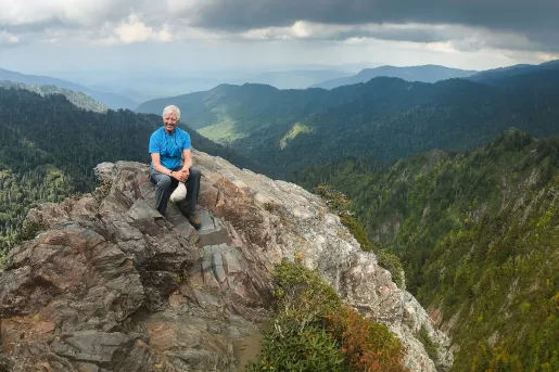 Guest sitting on rocky cliff-top. Overlooking forested valley.