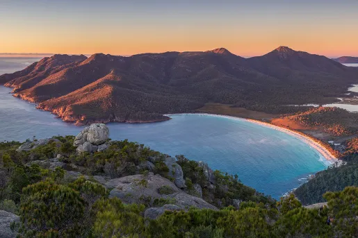 A panoramic landscape image of sunrise at the peak of Mount Amos with Wineglass Bay at the background, Freycinet National Park, Coles Bay, Tasmania, Australia.