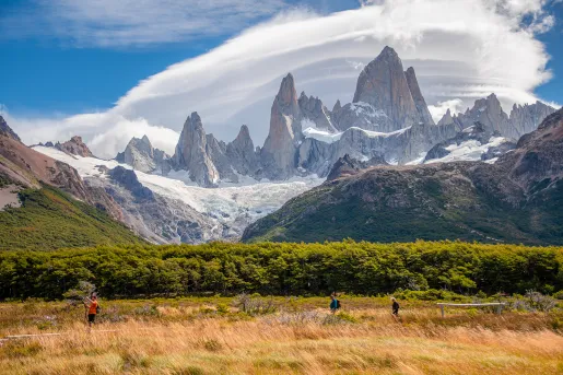 Three guests walking in grassland, forest, craggy mountains, clouds behind them.