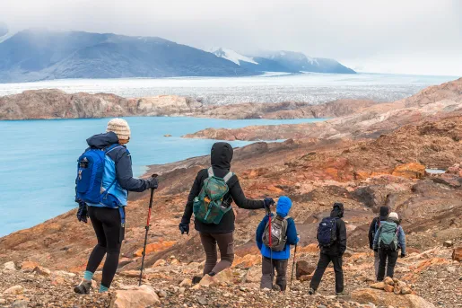 Six guests hiking down stony cliffside, blue water, mountains visible in distance.
