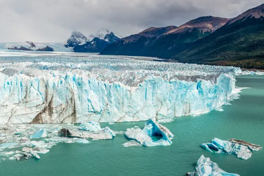 Wide shot of large glacial shelf, dark clouds and mountains in background.