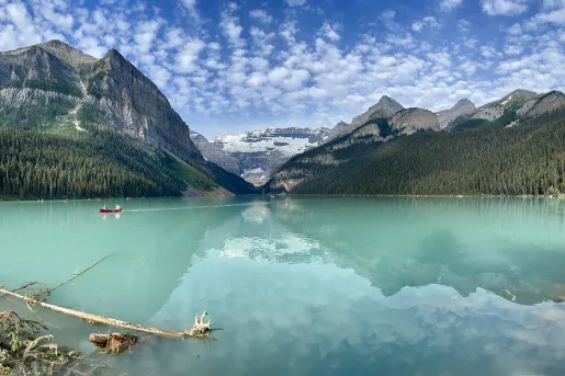 Wide shot of large blue lake, people canoeing in distance, mountains, clouds, sky in background.