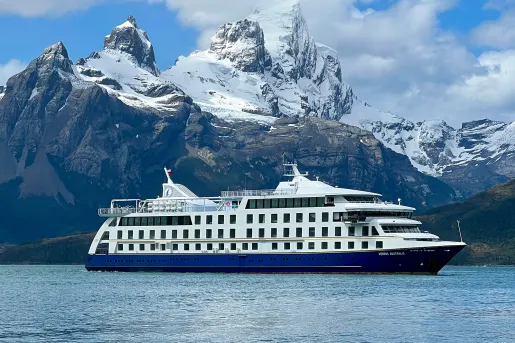 Wide shot of cruise ship, craggy mountains, clouds.