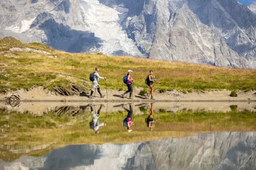 Three guests walking past reflective lake, meadow, snowy mountain in distance. 