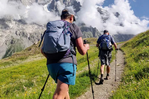 Two guests walking up mountain trail, clouds, mountain in distance. 
