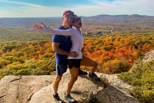 Two guests on clifftop, red/yellow/orange trees below.