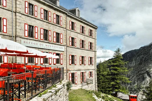 Stone hotel building exterior, with a metal railed balcony with red and it chairs and umbrellas