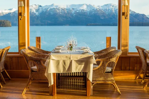 Outdoor dining table with woven chairs, a white table looking out towards a large windows to tall mountains