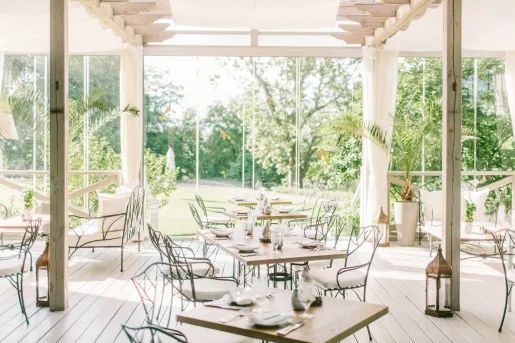 Indoor dining area with white chairs and wooden tables, with large glass walls surrounding