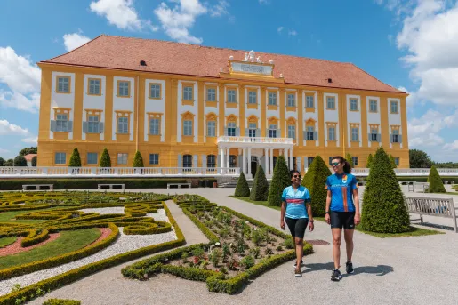 Two women laughing and walking in an outdoor garden in front of a yellow and white building