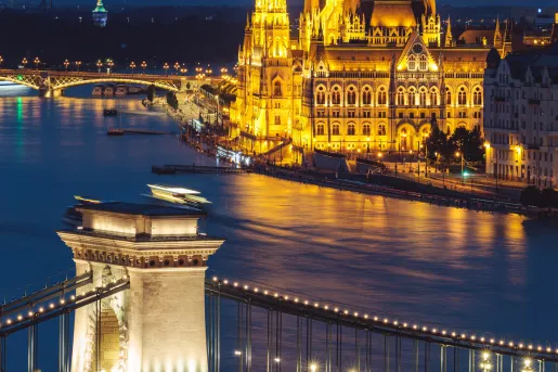 Illuminated castle-like building along the coast with a suspension bridge in front