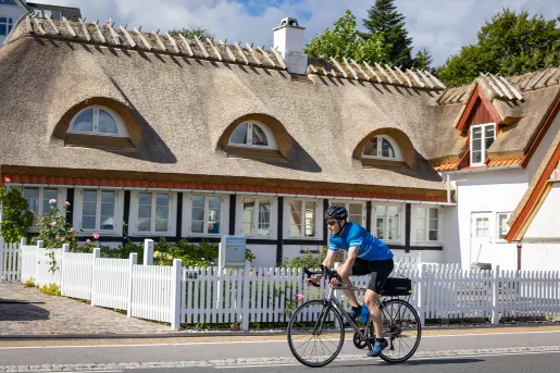 Man biking with a house and a white picket fence behind him