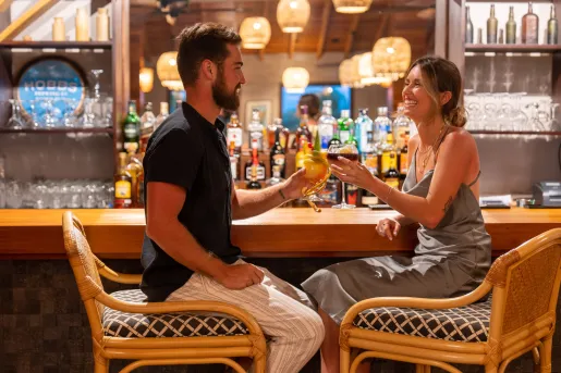 Man and woman sitting in front of a bar, raising glasses of beer and wine