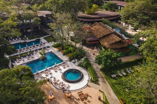 Sky view of outdoor pools and huts, with a modern building in the back