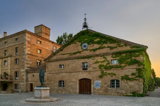 Exterior view of stone building covered in plants, with a bell on top