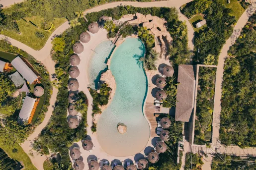 Sky view of outdoor pool surrounded by straw umbrellas and trees