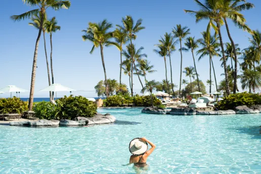 Woman wearing a hat swimming in a pool, surrounded by large palm trees