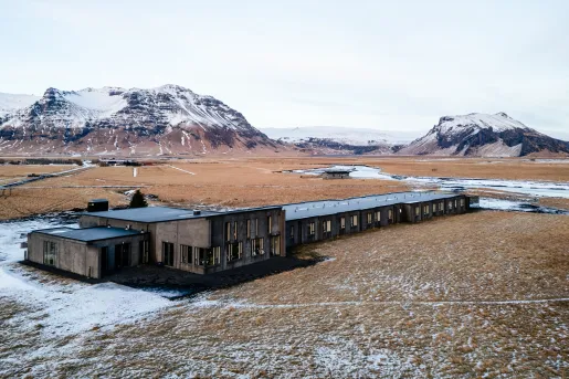 Exterior view of gray building in the middle of a valley, with snowy mountains in the background