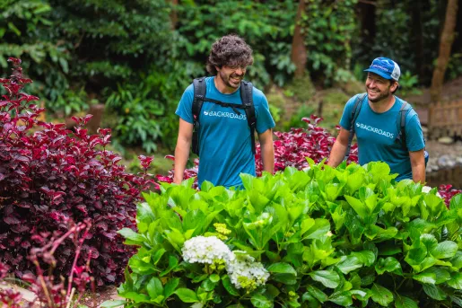 Two men walking through an outdoor path, surrounded by green and red plants