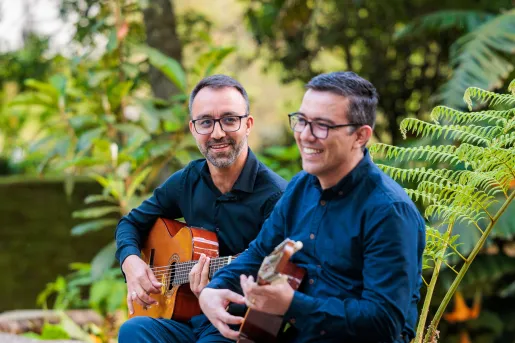 Two men sitting on a stone fence playing guitars and smiling