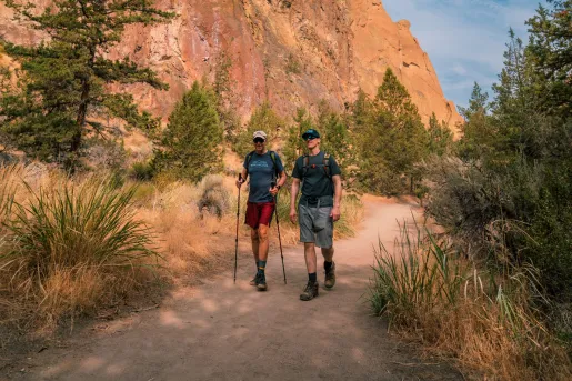Two men with walking poles, hiking on a dirt trail next to a canyon