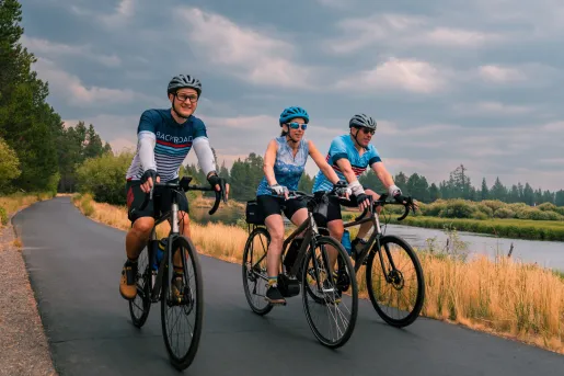 Two men and one woman riding bicycles on an asphalt road, next to a river