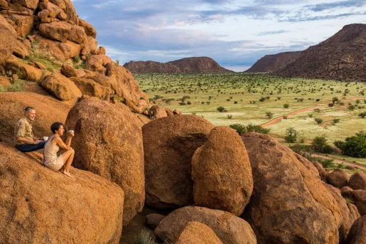 Man and woman sitting on a large boulder overlooking a large, grassy valley