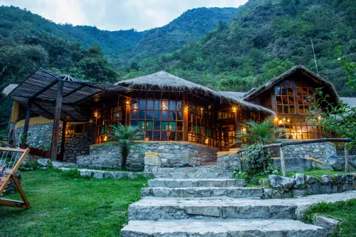 Exterior view of stone building with straw hut roof, with tree-covered mountains in the background