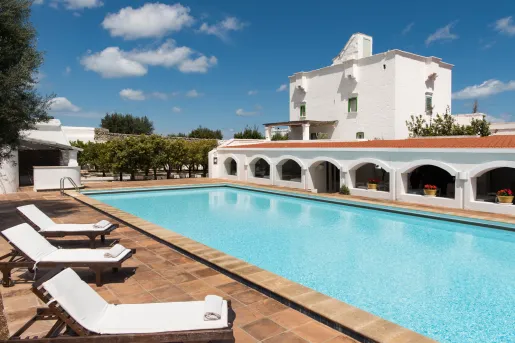 Outdoor pool, surrounded by white pool chairs and a white, rectangular building