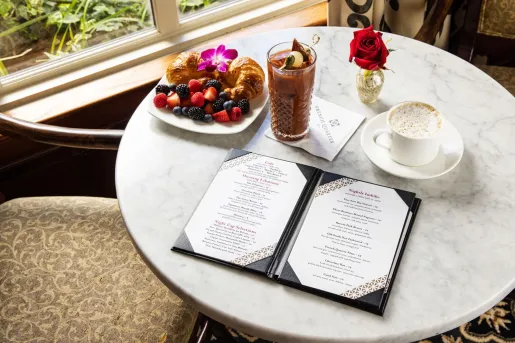 Circular, marble table with an open menu, a cup of coffee and a plate of pastries and fruits