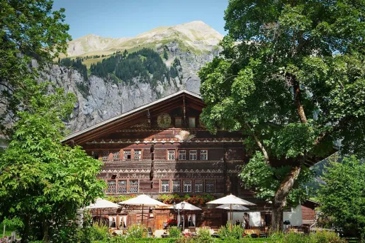 Exterior view of a wooden-style cabin, surrounded by trees and a tall mountain in the background