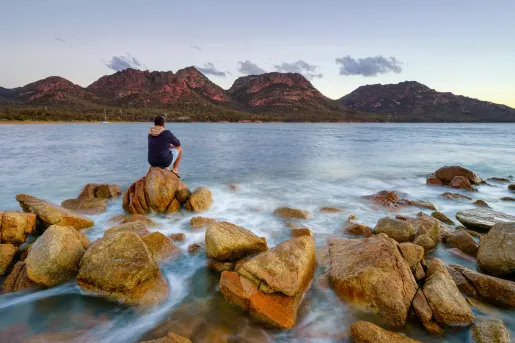 Man sitting on a boulder by the oceanside, looking out to large cliffs