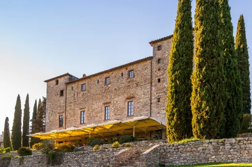 Tall stone building with tall trees and an outdoor patio covered by yellow umbrellas