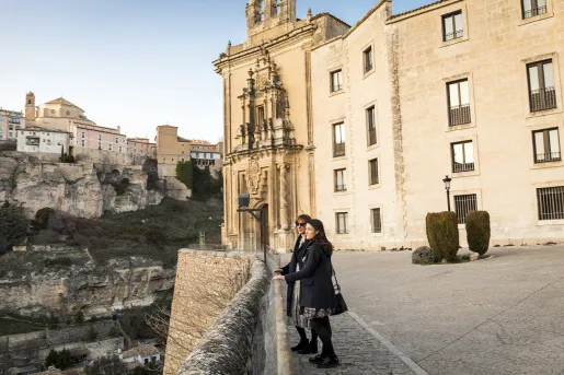 Two women standing on a stone barricade next to a cliff, looking out to other buildings