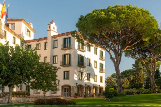 Exterior view of white and stone hotel building, surrounded by tall trees and a grass field