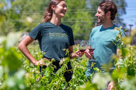 Man and woman smiling while holding up wine glasses, standing in a vineyard