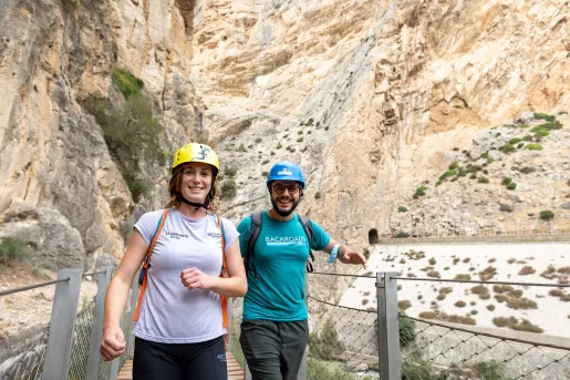 Man and woman walking on a wooden bridge with a large cliff in the background