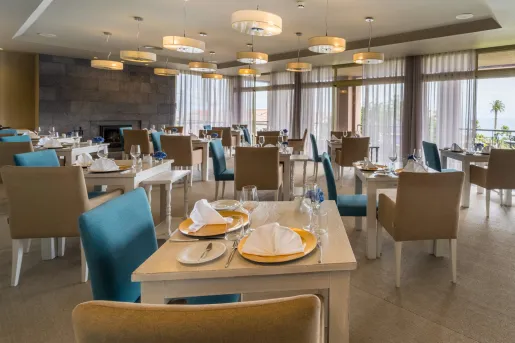 Restaurant dining hall with blue and beige cushioned chairs and large curtains along the windows