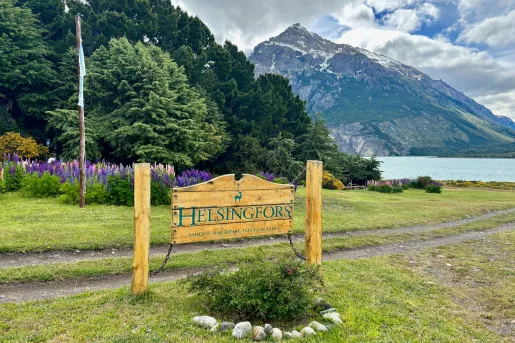 Wooden sign with the word "Helsingfors", in front of tall trees and an open lake