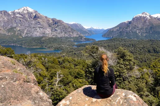 Woman sitting on a large boulder, looking to a valley of trees and a lake in the distance