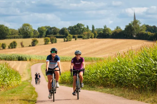 Two women riding bikes on a road, surrounded by fields of crops