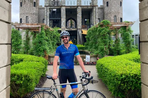 Man standing behind his bike, smiling with a large, stone building in the background