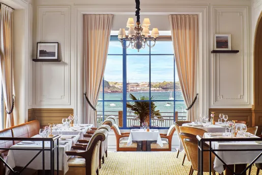 Restaurant dining hall with white chairs and tables, with large windows looking out to the ocean
