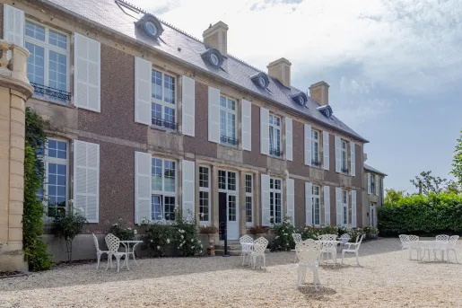 Exterior view of beige, stone building with a courtyard with white tables and chairs in front