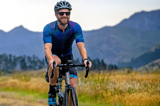 Man wearing biking gear, riding a bicycle on an asphalt road with tall mountains in the background