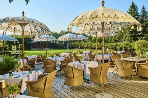 Outdoor patio with dining tables and woven chairs, with decorated umbrellas and a grass field in the background