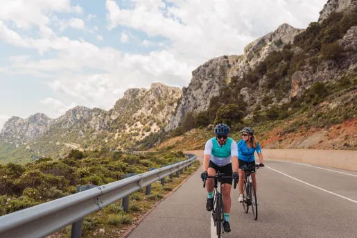 Man and woman riding bikes on an empty road, with large cliffs in the background
