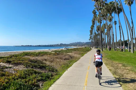 Woman riding bike on a road with palm trees on the right and ocean on the left