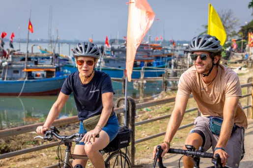 Man and woman riding bikes on a road next to a dock, with floating boats in the water